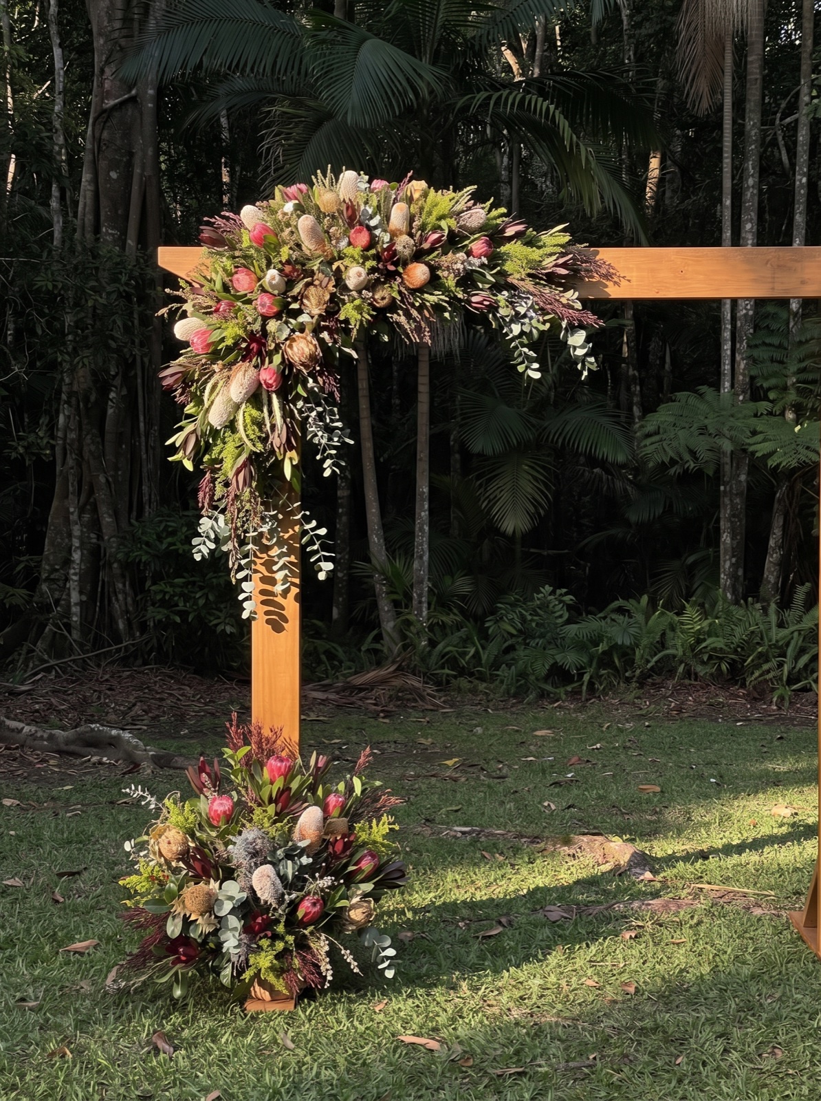 Botanical flowers on the upper corner of a timber ceremony arch, with trailing greenery and a matching arrangement at its base in a garden setting.