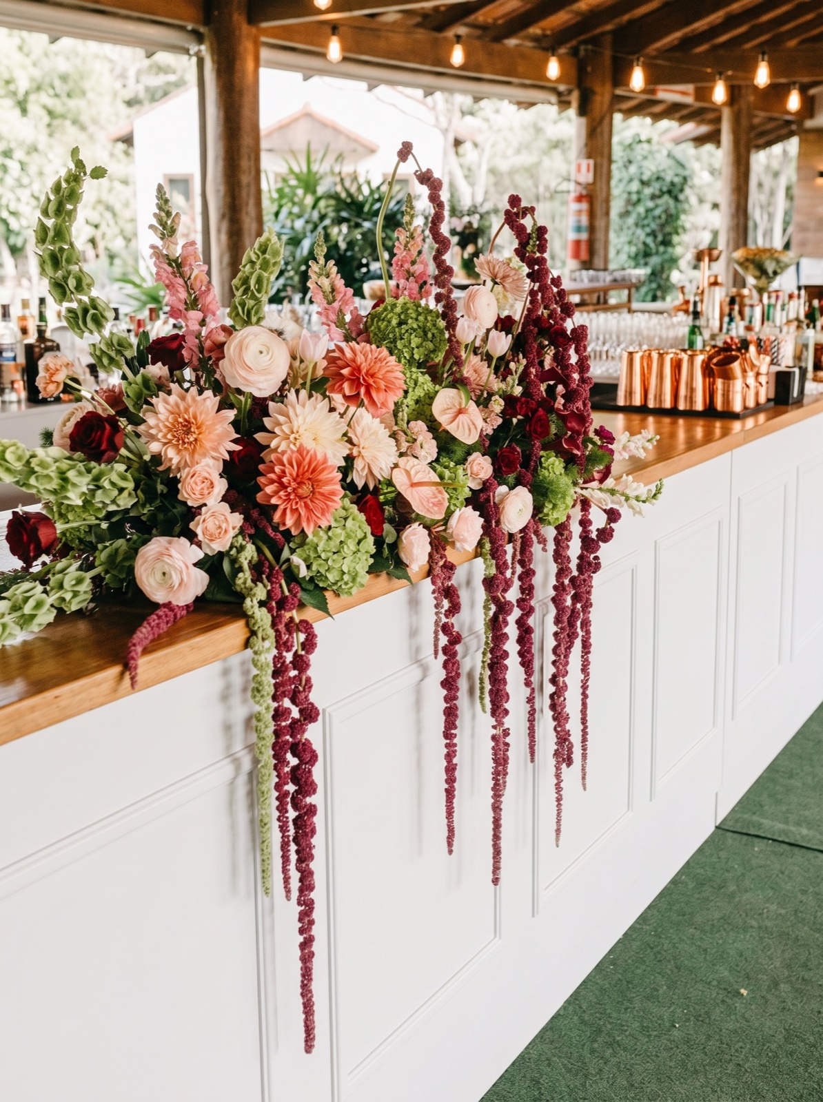 A lush, broad blush, coral, burgundy and green installation across a timber bar, with long burgundy trails over its white front.