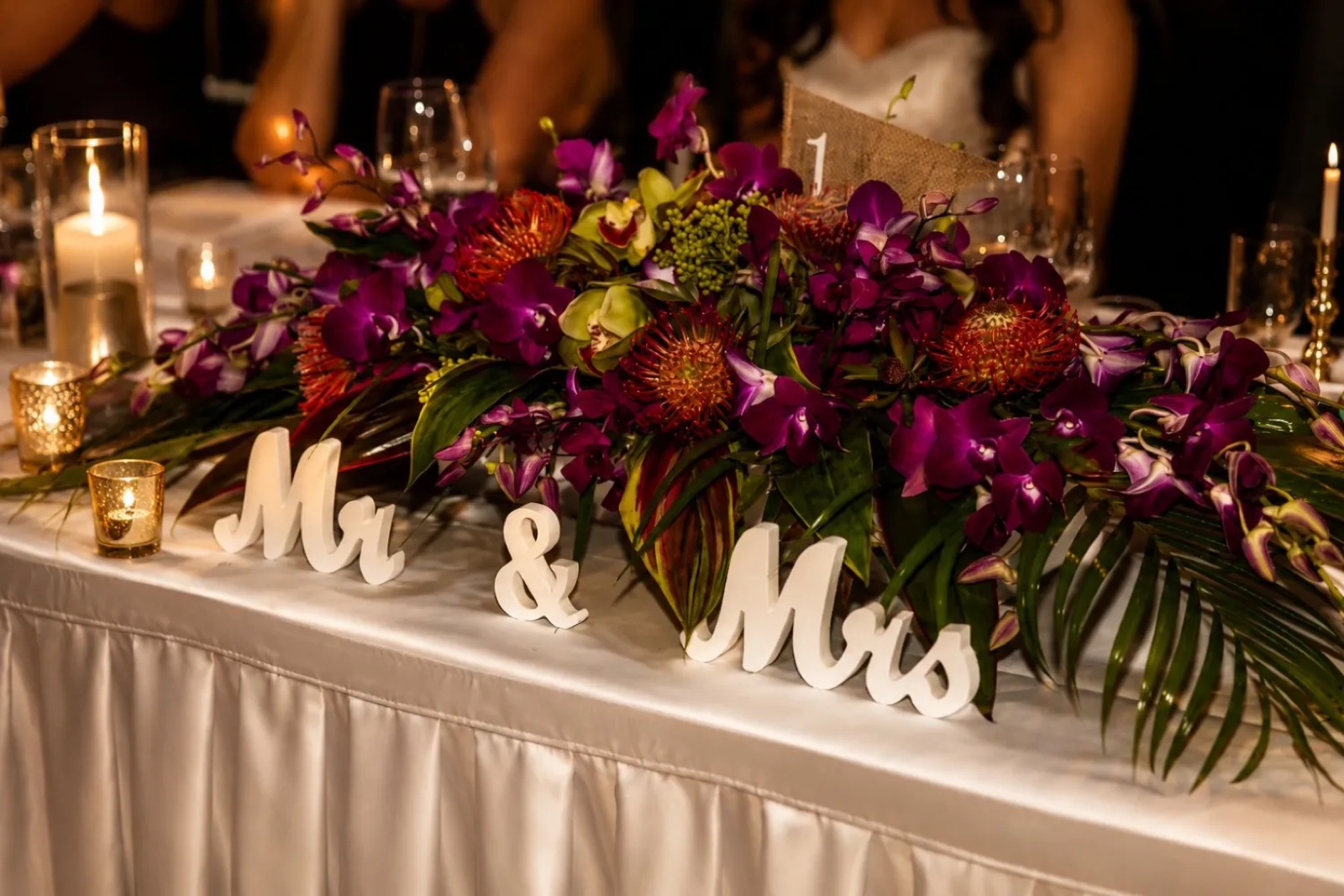 Low, broad wedding table flowers in purple, orange and green, with candles and a white tablecloth.
