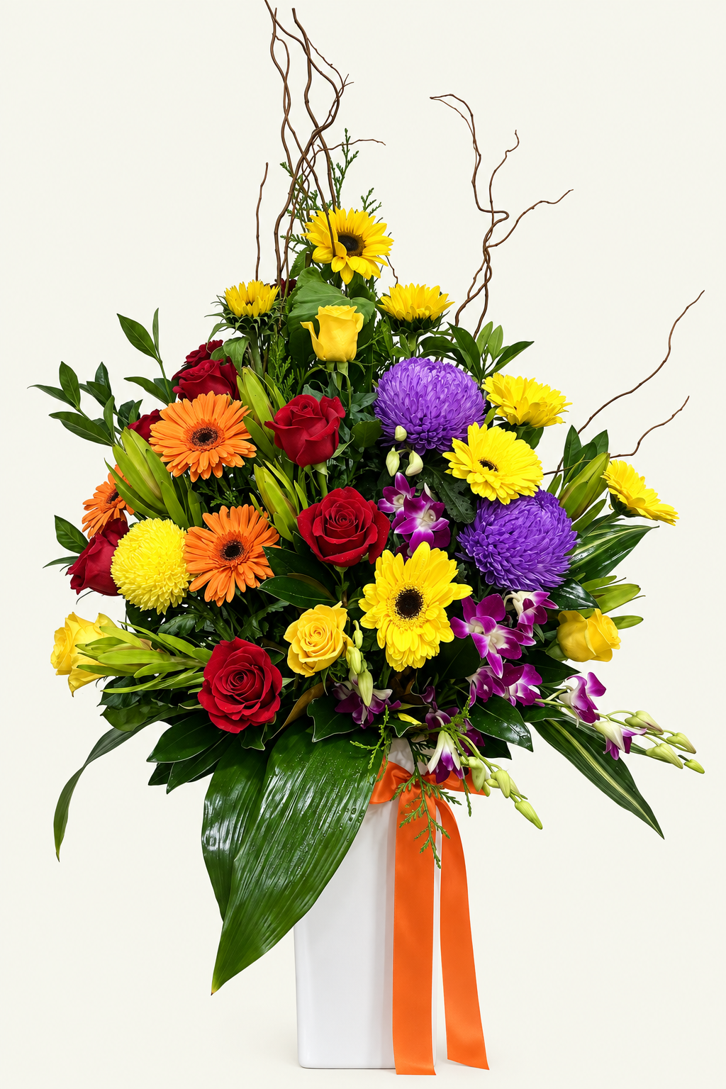 Colourful arrangement of sunflowers, red and yellow roses, orange gerberas, purple flowers and green foliage in a white vase with an orange ribbon